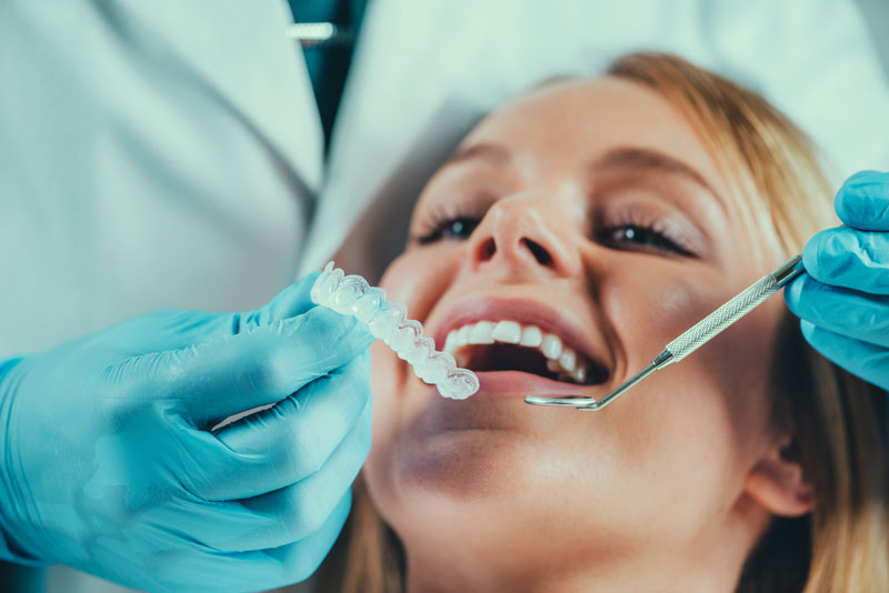 A dentist, wearing blue gloves, is holding a clear dental aligner and is about to place or adjust it in the mouth of a female patient, who appears relaxed and has her mouth open.