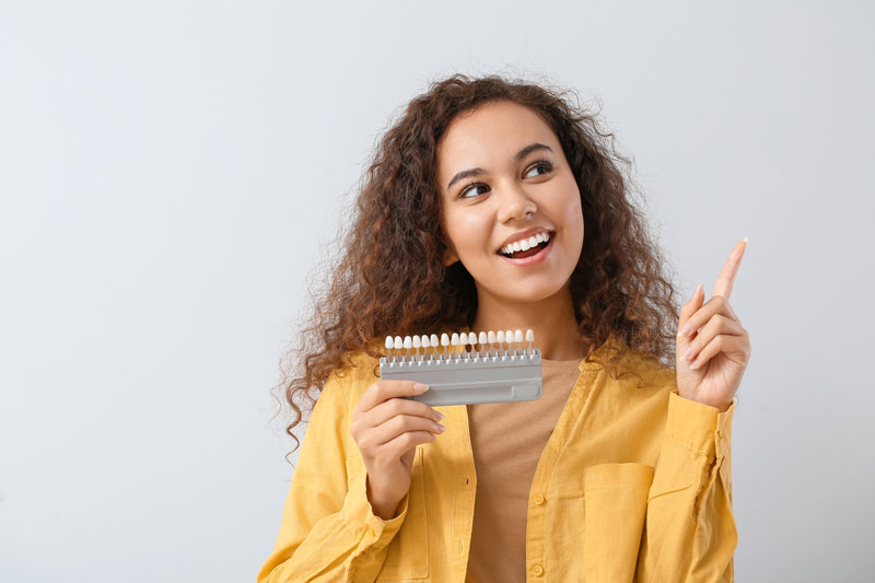 Young woman smiling, holding a dental veneers sample, wearing a yellow jacket, showcasing potential dental cosmetic improvements.