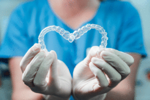 Female doctor holding two transparent heart-shaped dental aligners, dental office in the background, blurred.