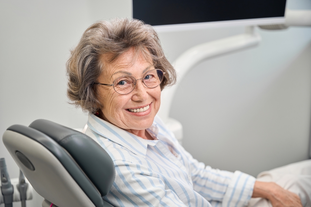 Photo of a dentist smiling and pointing to a digital screen that shows a detailed cost breakdown of dental implant procedures, with line items for the implant, abutment, crown, and other fees. The text on the screen reads "Understanding Your Implants Teeth Price".